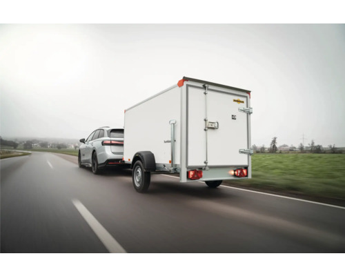 Une remorque fourgon Humbaur blanche avec des portes arrière est tractée par une voiture sur une route de campagne.
