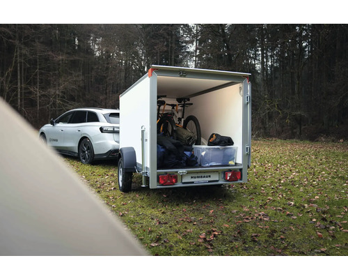 Remorque fourgon Humbaur blanche attelée à une voiture, chargée d'un VTT et de matériel dans une forêt.