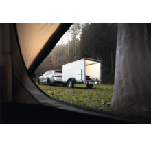 Voiture argentée avec une remorque fourgon blanche dans un champ devant une forêt, vue depuis l'intérieur d'une tente.