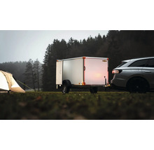 Remorque fourgon Humbaur attelée à une voiture sur une prairie à côté d'une tente éclairée devant une forêt.
