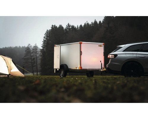 Remorque fourgon Humbaur attelée à une voiture sur une prairie à côté d'une tente éclairée devant une forêt.