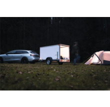 Voiture argentée avec remorque fermée blanche sur un camping dans la forêt la nuit. La remorque est éclairée et une tente se trouve à côté.