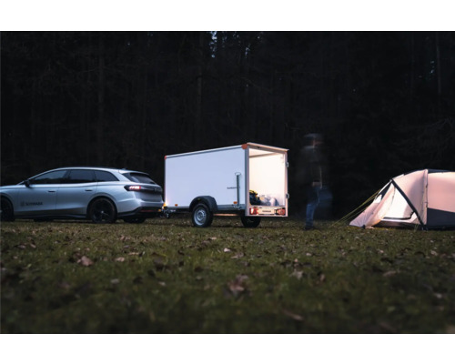 Voiture argentée avec remorque fermée blanche sur un camping dans la forêt la nuit. La remorque est éclairée et une tente se trouve à côté.