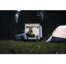 Remorque fourgon Humbaur avec portes ouvertes, chargée d'un vélo et de matériel de camping, à côté d'une tente dans une clairière.