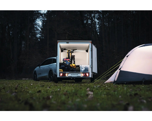 Remorque fourgon Humbaur avec portes ouvertes, chargée d'un vélo et de matériel de camping, à côté d'une tente dans une clairière.