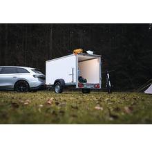 Remorque fourgon blanche Humbaur attelée à une voiture en forêt, avec porte arrière ouverte, éclairage intérieur et matériel de camping.