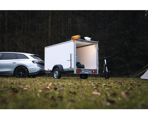 Remorque fourgon blanche Humbaur attelée à une voiture en forêt, avec porte arrière ouverte, éclairage intérieur et matériel de camping.