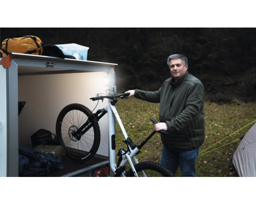 Un homme se tient à l extérieur à côté d une remorque fermée éclairée et tient un vélo tout terrain blanc.