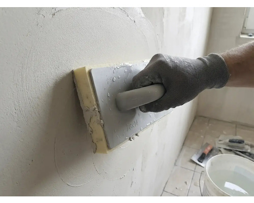 Main gantée utilisant une taloche éponge pour lisser de l'enduit sur un mur.