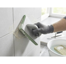 Une personne jointoyant des carreaux muraux avec une taloche à joints. Seau d'eau avec éponge et truelle en arrière-plan.