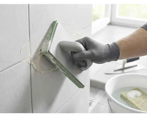 Une personne jointoyant des carreaux muraux avec une taloche à joints. Seau d'eau avec éponge et truelle en arrière-plan.