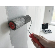 Eine Hand trägt mit einer Farbwalze mit rotem Griff graue Farbe auf eine Wand auf. Ein Werkzeugkoffer ist im Hintergrund zu sehen.
