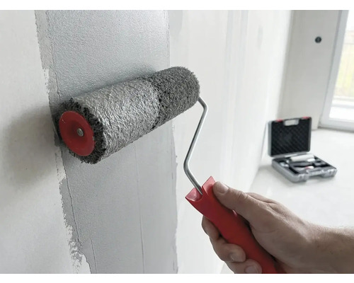 Eine Hand trägt mit einer Farbwalze mit rotem Griff graue Farbe auf eine Wand auf. Ein Werkzeugkoffer ist im Hintergrund zu sehen.