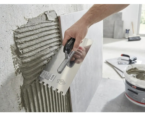 Eine Hand trägt mit einer Glättkelle mit Zahnung Fliesenkleber auf eine Wand auf.