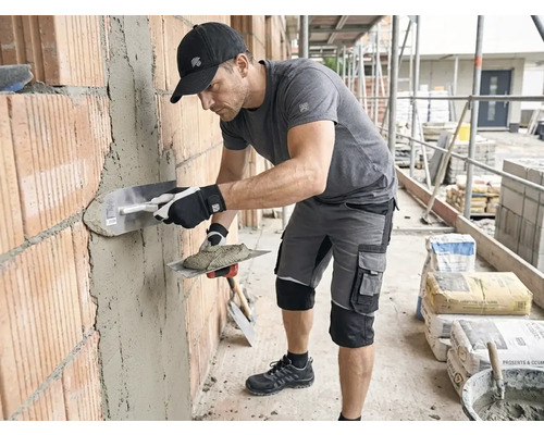 Handwerker in Arbeitskleidung verputzt eine Ziegelwand auf einer Baustelle mit einer Glättkelle und Mörtel.