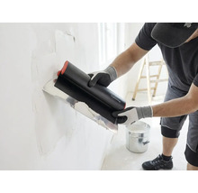 Handwerker trägt mit einem breiten Flächenspachtel Spachtelmasse auf eine Wand auf, im hellen Raum stehen ein Eimer und eine Leiter.