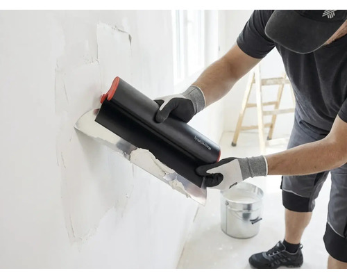 Handwerker trägt mit einem breiten Flächenspachtel Spachtelmasse auf eine Wand auf, im hellen Raum stehen ein Eimer und eine Leiter.