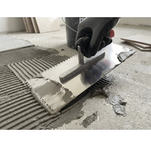 Eine Person trägt mit einer metallenen Zahnkelle Fliesenkleber auf einen Betonboden auf.