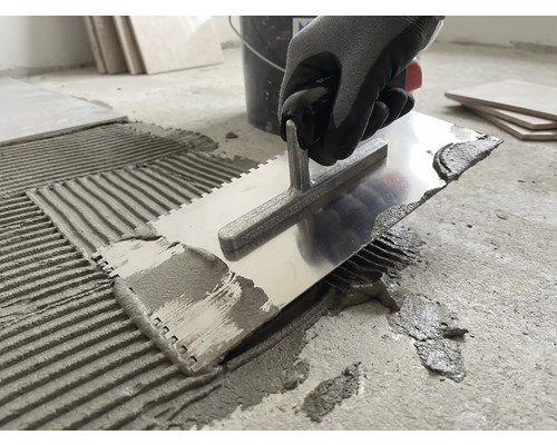 Eine Person trägt mit einer metallenen Zahnkelle Fliesenkleber auf einen Betonboden auf.