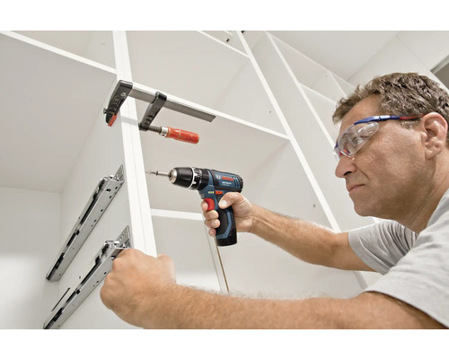 Ein Mann mit Schutzbrille montiert Schubladenschienen in einem weißen Schrank mit einem Bosch Akku-Bohrschrauber und einer Schraubzwinge.