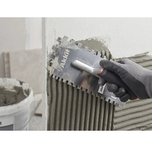 Une main gantée applique uniformément de la colle à carrelage sur un mur avec une taloche crantée Akkit.