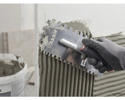 Une main gantée applique uniformément de la colle à carrelage sur un mur avec une taloche crantée Akkit.