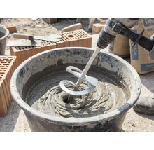 Un mélangeur sur une perceuse mélange du mortier gris dans un seau sur un chantier avec des briques et des truelles.