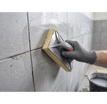 Eine Hand reinigt frisch verfugte Wandfliesen mit einem Schwammbrett und Wasser.