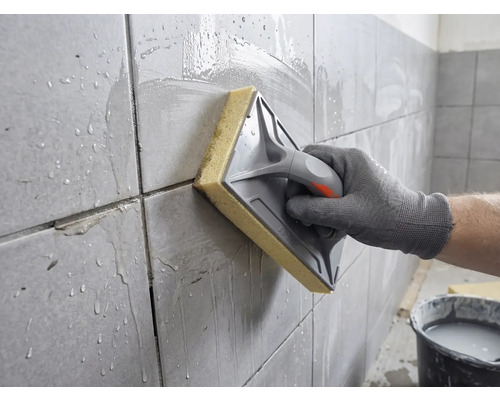 Eine Hand reinigt frisch verfugte Wandfliesen mit einem Schwammbrett und Wasser.