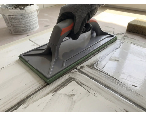 Main avec gant de protection utilisant un platoir à jointer pour jointoyer des carreaux.