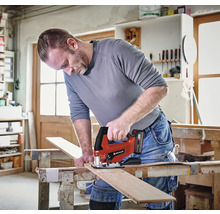 Un artisan utilise une scie sauteuse Einhell sans fil pour couper une planche en bois sur un établi dans un atelier lumineux. Einhell Logo.