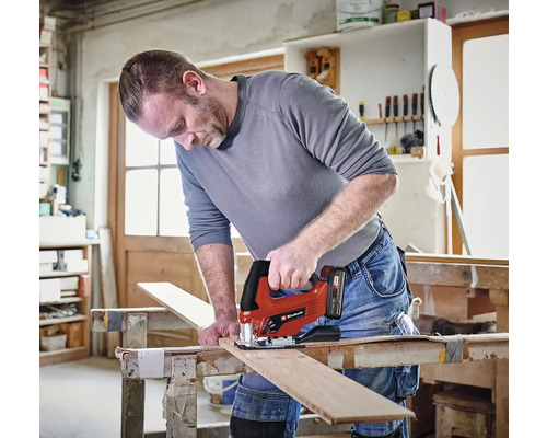 Un artisan utilise une scie sauteuse Einhell sans fil pour couper une planche en bois sur un établi dans un atelier lumineux. Einhell Logo.