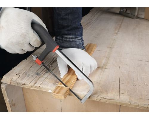 Person mit weissen Schutzhandschuhen schneidet eine Holzleiste mit einer kleinen Handsäge auf einer hölzernen Werkbank.