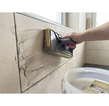 Eine Person reinigt beigefarbene Wandfliesen mit einem Schwammbrett. Ein weisser Eimer mit Wasser steht im Vordergrund.