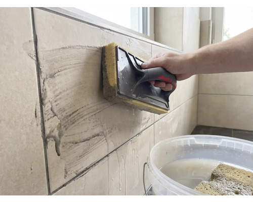 Eine Person reinigt beigefarbene Wandfliesen mit einem Schwammbrett. Ein weisser Eimer mit Wasser steht im Vordergrund.