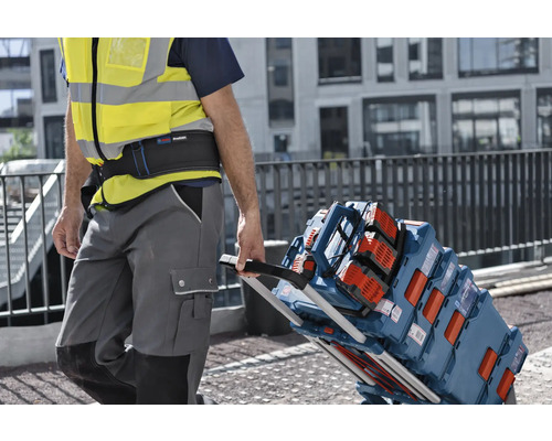 Handwerker mit Warnweste zieht eine Sackkarre beladen mit Bosch L-BOXX Werkzeugkoffern und einem Baustellenradio auf einer Baustelle.