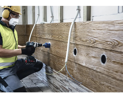 Un artisan perce une ouverture dans un revêtement mural à l'aspect bois avec une perceuse-visseuse sans fil et une scie cloche.