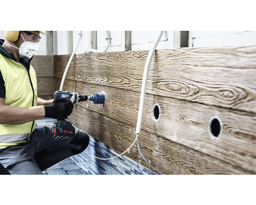 Artisan utilisant une perceuse-visseuse sans fil avec scie cloche pour percer un panneau mural avec aspect bois sur un chantier.
