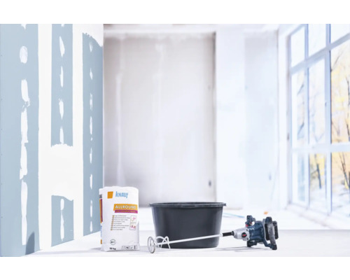 Un sac de 20 kilogrammes d'enduit Knauf Allround à côté d'un seau à mélanger noir et d'un malaxeur électrique sur un chantier lumineux.