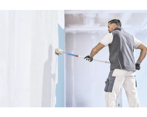 Artisan en vêtements de travail peignant un mur avec un rouleau à peinture et une perche télescopique dans un intérieur lumineux.