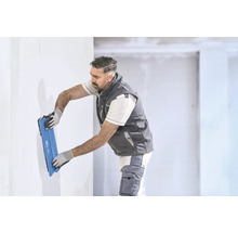 Un artisan lisse un mur blanc avec un large couteau à enduire bleu dans une pièce lumineuse.