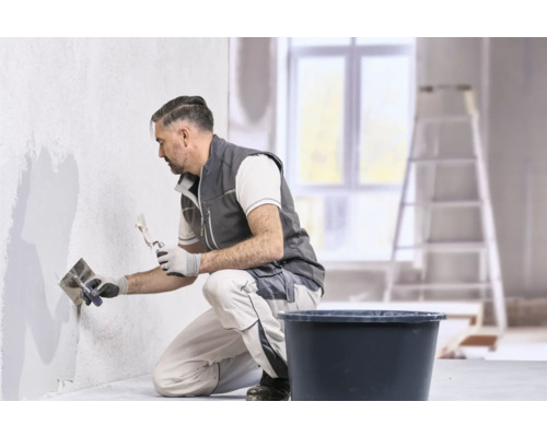 Un artisan en vêtements de travail applique de l'enduit sur un mur avec une lisseuse et un couteau à enduire.