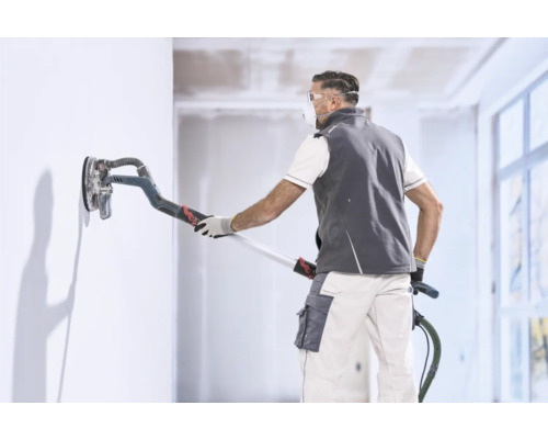 Un artisan lisse un mur avec une ponceuse girafe dans une pièce intérieure lumineuse.