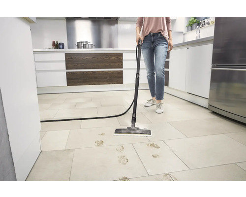 Personne nettoyant des traces de pas boueuses sur des dalles de sol claires dans une cuisine moderne et lumineuse avec un nettoyeur vapeur.