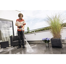 Un homme nettoie les dalles de la terrasse avec un nettoyeur haute pression. Des meubles de jardin et des bacs a fleurs sont visibles.