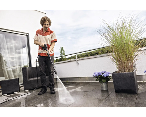 Un homme nettoie les dalles de la terrasse avec un nettoyeur haute pression. Des meubles de jardin et des bacs a fleurs sont visibles.