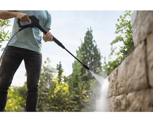 Une personne nettoie un mur en pierre structuré avec la lance d'un nettoyeur haute pression dans un jardin.