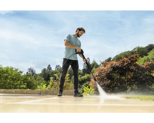 Un homme nettoie une terrasse en pierre claire dans le jardin avec un nettoyeur haute pression par temps ensoleillé.