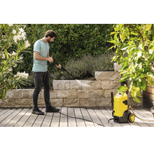 Un homme nettoie un mur en pierre dans un jardin avec un nettoyeur haute pression jaune sur une terrasse en bois entourée de plantes.
