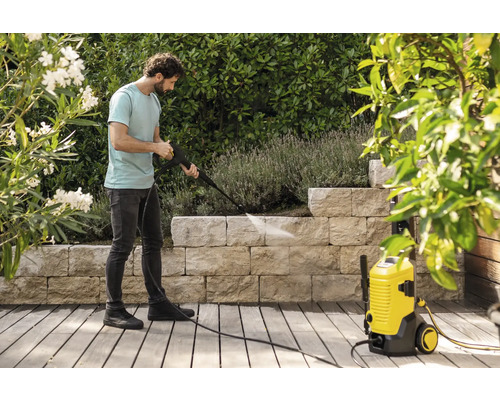 Un homme nettoie un mur en pierre dans un jardin avec un nettoyeur haute pression jaune sur une terrasse en bois entourée de plantes.
